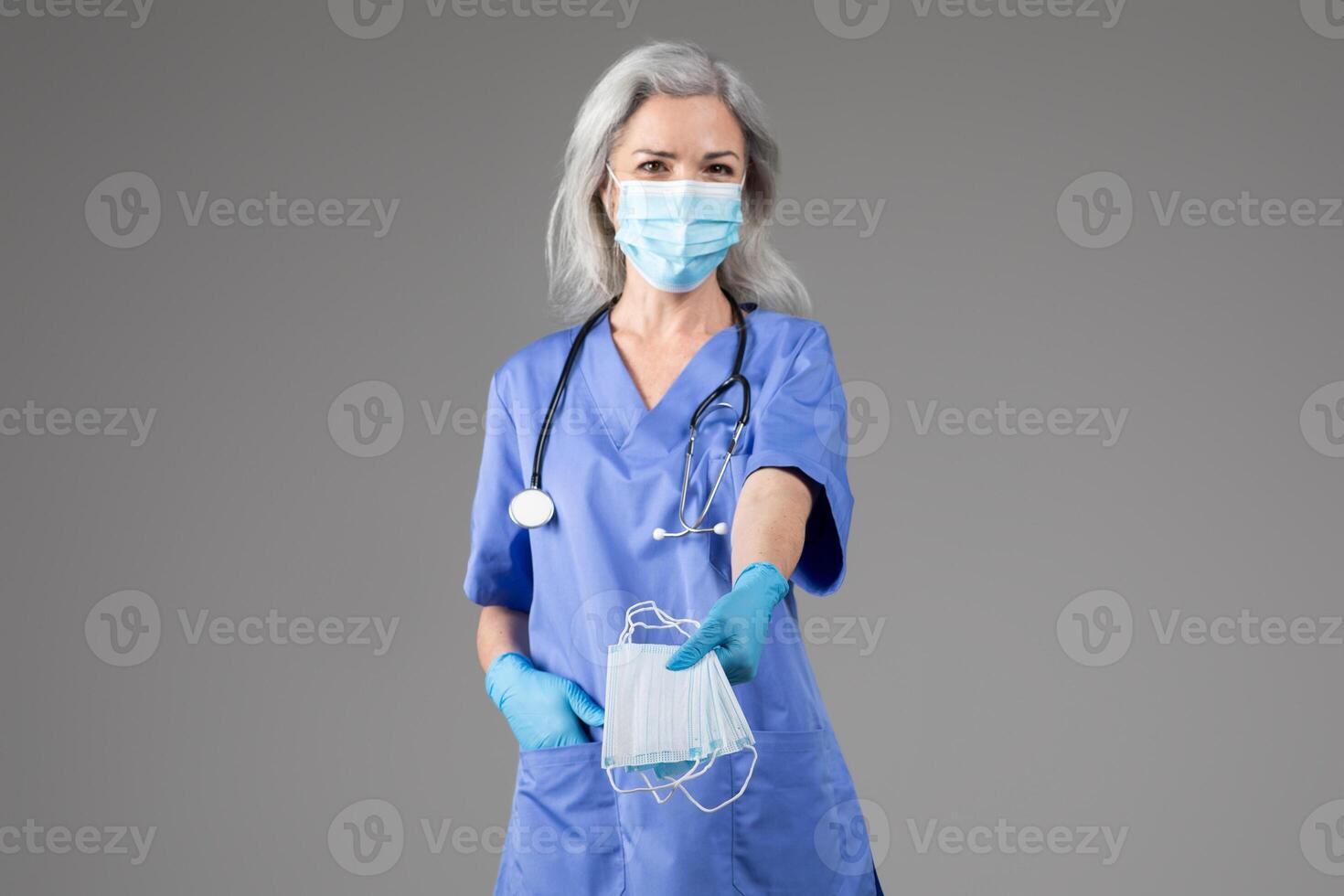 A middle aged woman in scrubs is presenting safety masks with gloved hands. Her calm demeanor underscores the importance of health precautions in a medical environment during crucial times. photo