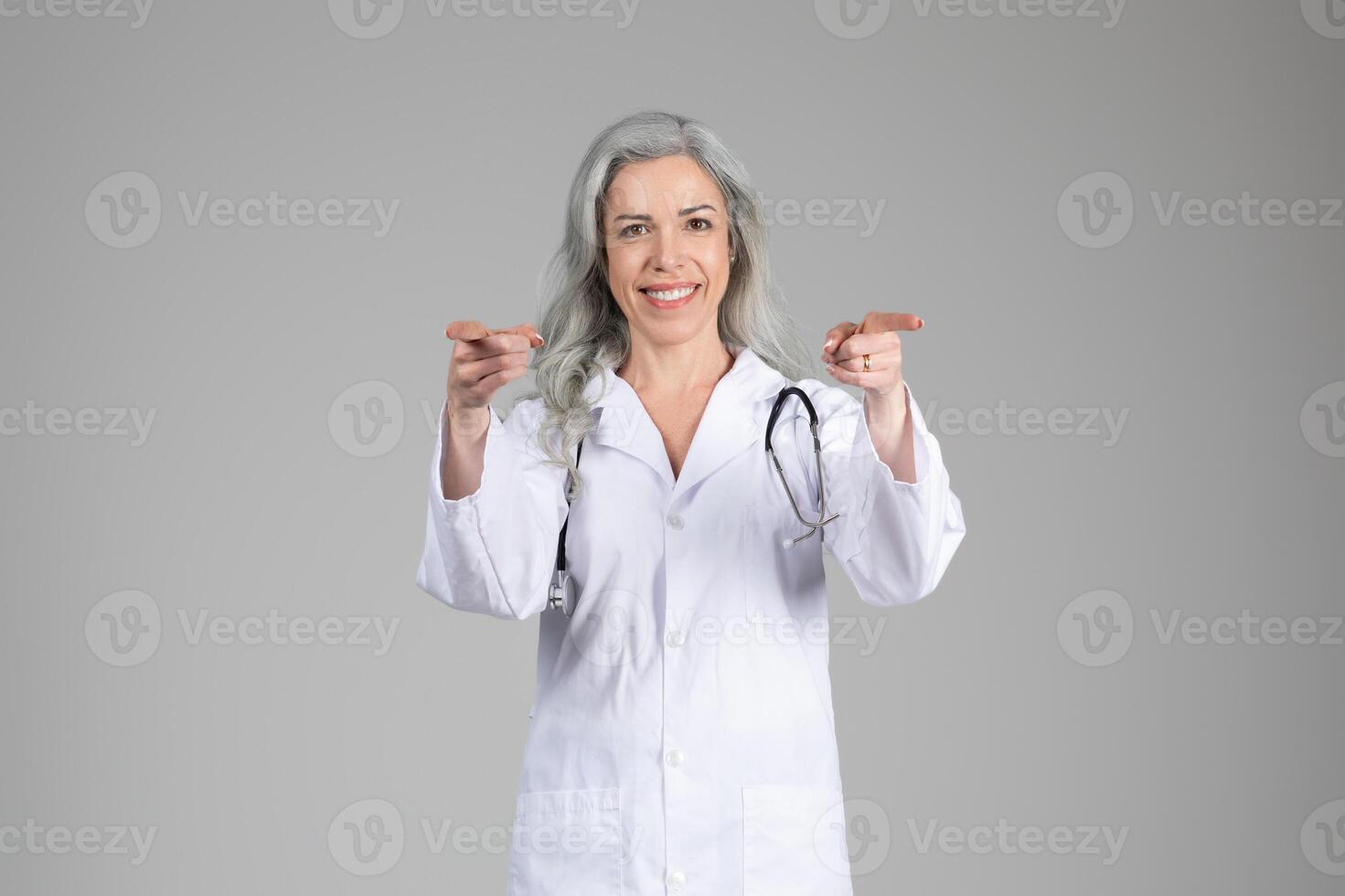 A mature woman in a white lab coat stands confidently, smiling while pointing toward the viewer. She has long gray hair and a stethoscope around her neck, indicating her role in healthcare. photo