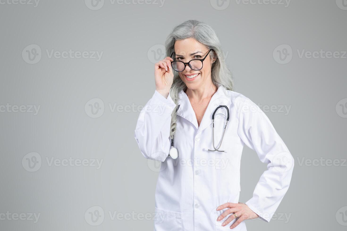 A middle aged woman doctor wearing a lab coat and glasses is smiling confidently. She stands with her hand on her hip, radiating a positive demeanor in a neutral-toned setting, copy space photo