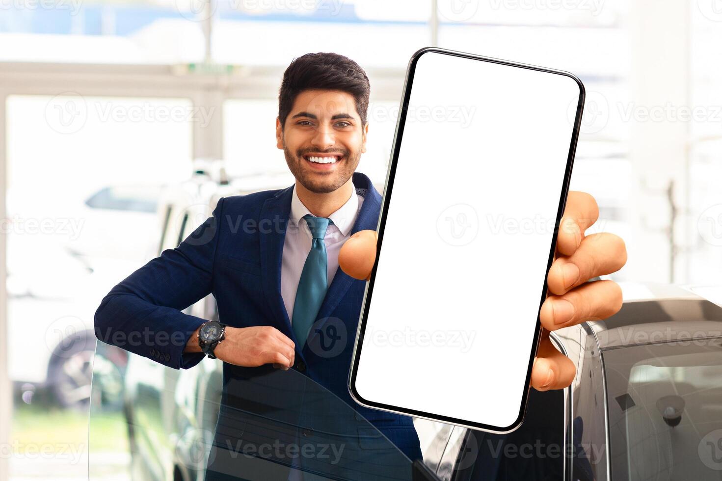 Emotional middle eastern businessman choosing new car, standing by brand new black auto, showing mobile phone with white blank screen and smiling, using auto dealership app, mockup photo