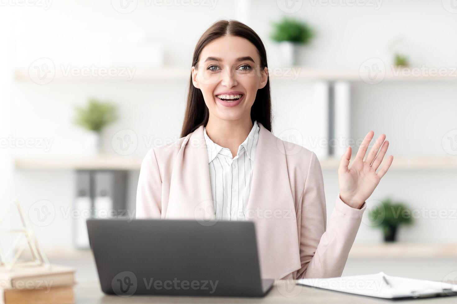 Cheerful Female Manager Waving Hello Sitting At Laptop And Smiling To Camera Consulting Clients Online In Modern Office Indoor. Travel Agent Having Distance Consultation Concept photo