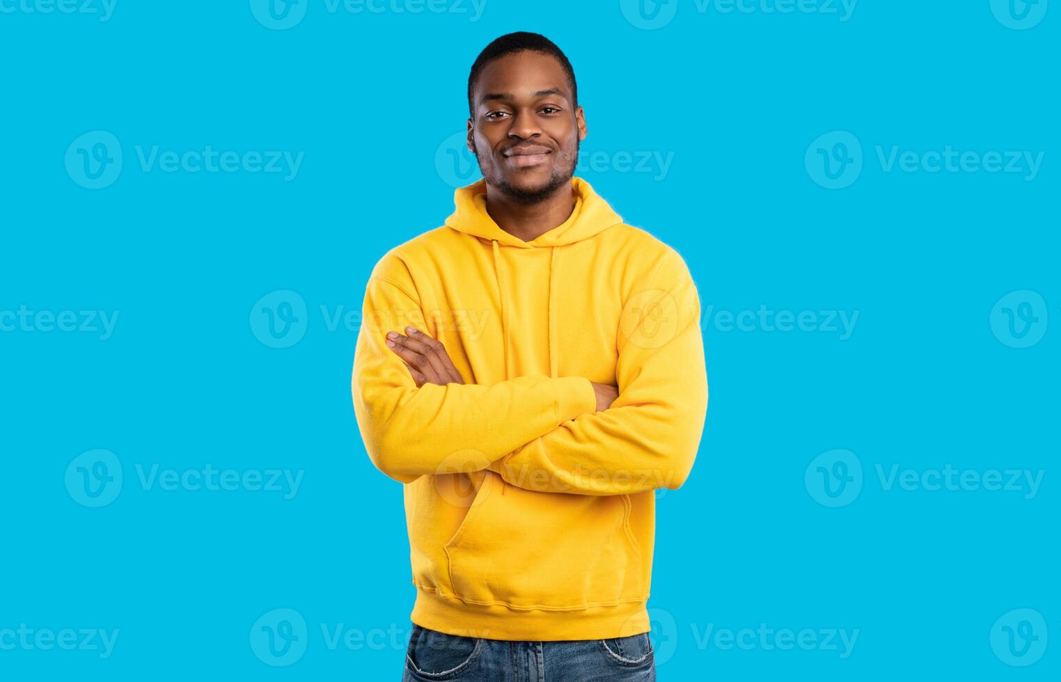 Handsome Black Millennial Man Crossing Hands Posing Smiling To Camera Standing In Studio On Blue Background, Wearing Yellow Hoodie. Shot Of Confident African American Guy photo