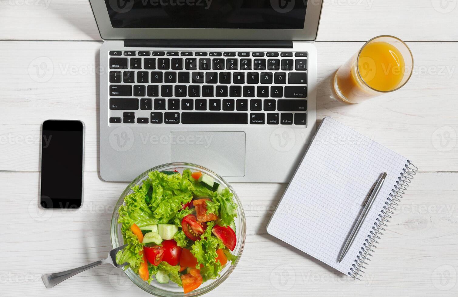 Healthy business lunch in the office, top view of vegetable salad on white wooden desk near laptop computer keyboard. Salad bowl, juice, mobile phone and notepad with pen flat lay. Snack at break time photo