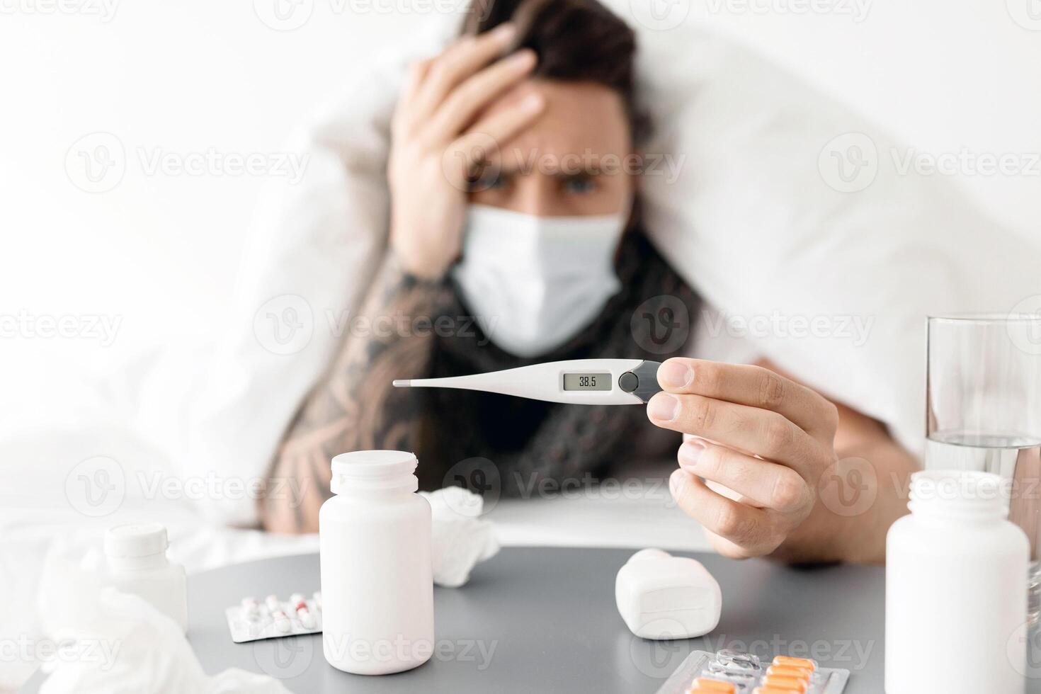 Ill young man lying under blanket on bed and showing thermometer with temperature result to camera. A lot of pills on table. Covid-19 coronavirus disease, seasonal flu and cold, illness fatigue photo