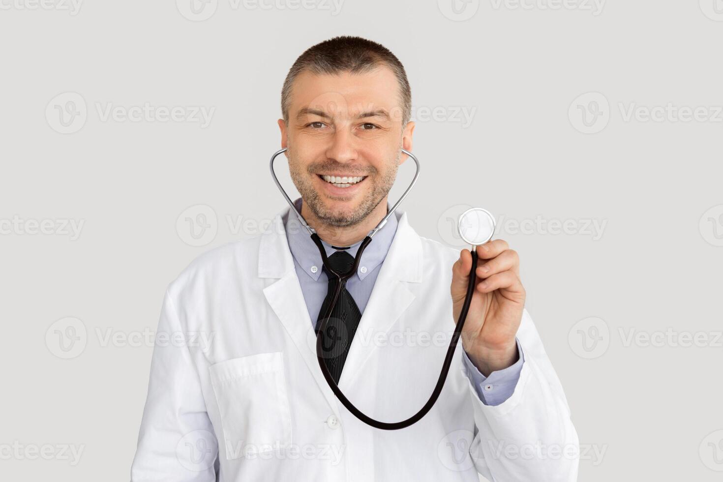 Medical Checkup Concept. Portrait of smiling confident mature general practitioner holding stethoscope, ready to examine patient, standing isolated over white studio background, looking at camera photo