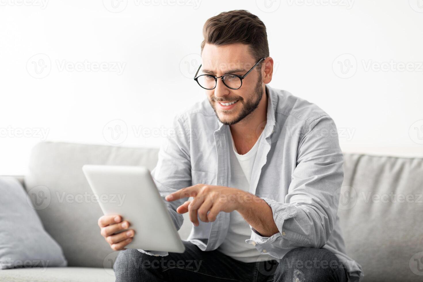 Free time concept. Happy man using digital tablet, sitting on couch in living room, copy space. Cheerful guy checking social media or chatting with friends on pad photo
