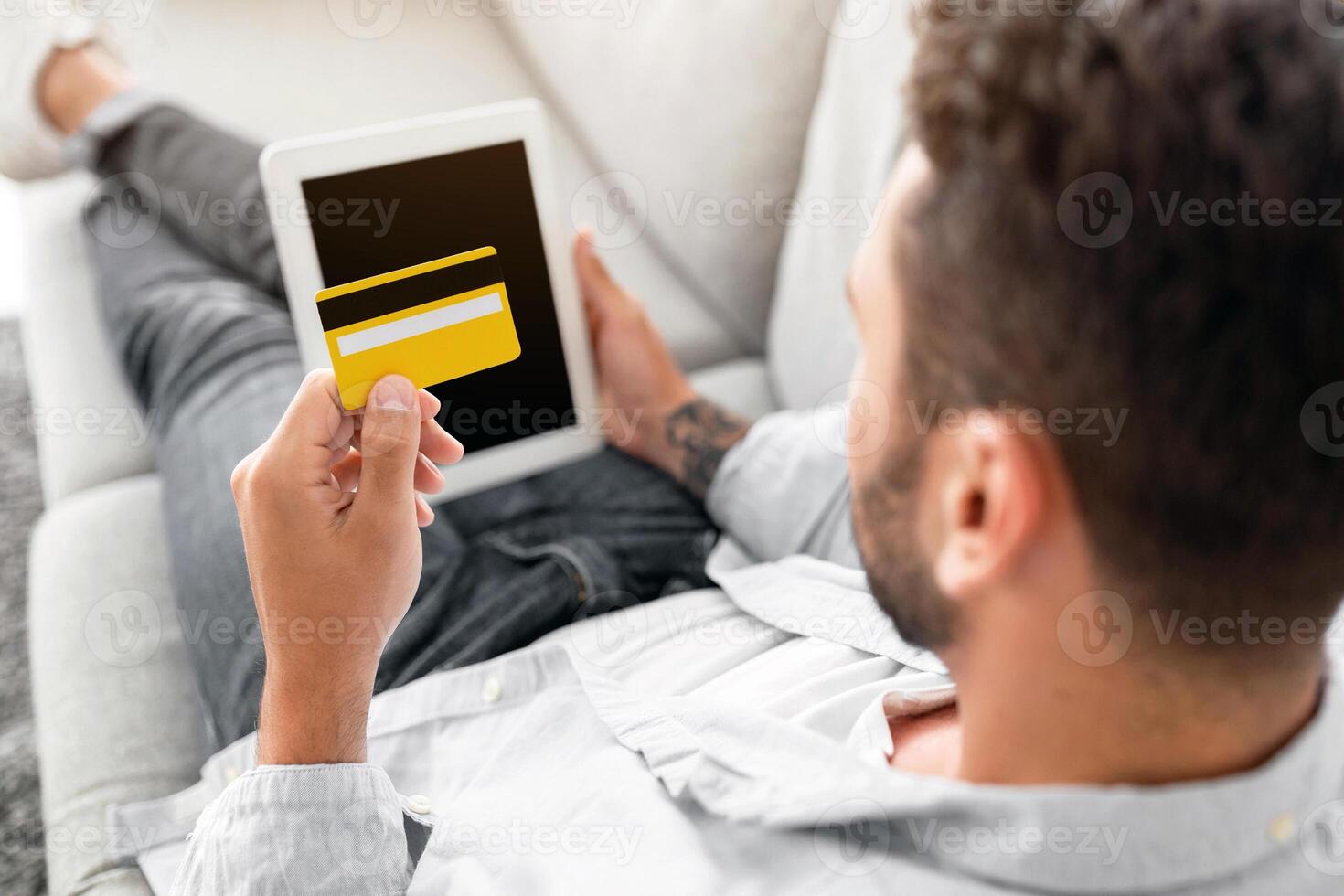 E-commerce concept. Unrecognizable man with credit card and digital tablet sitting on sofa at home, over shoulder shot. Young guy shopping online, using modern pad and holding card photo