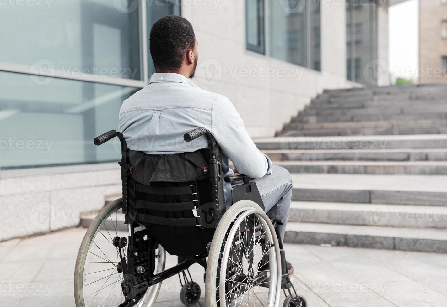 Back view of young black guy in wheelchair standing in front of stairs without ramp, having problem entering building, empty space. Impairment and accessible city environment concept photo