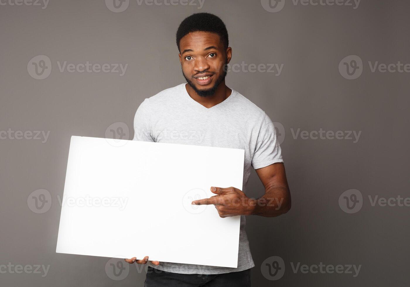 Picture of young smiling african-american man holding white blank board and pointing on it, on grey background, copy space photo