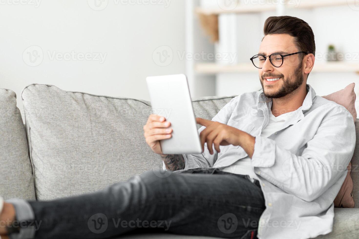 Technology concept. Smiling millennial man using digital tablet resting on couch at his modern apartment, free space. Happy guy browsing internet on pad, resting on sofa in living room photo