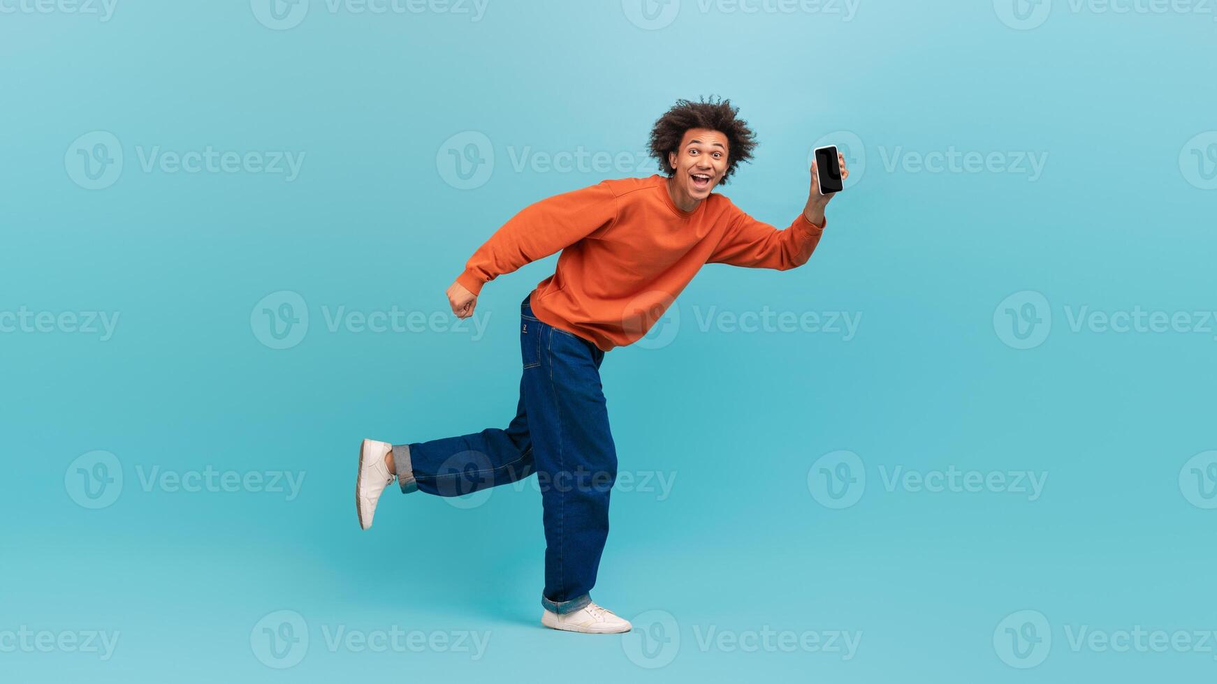 A young African American man with curly hair is running happily against a light blue background. He wears a bright orange sweater and blue jeans, holding a smartphone as he enjoys his moment. photo