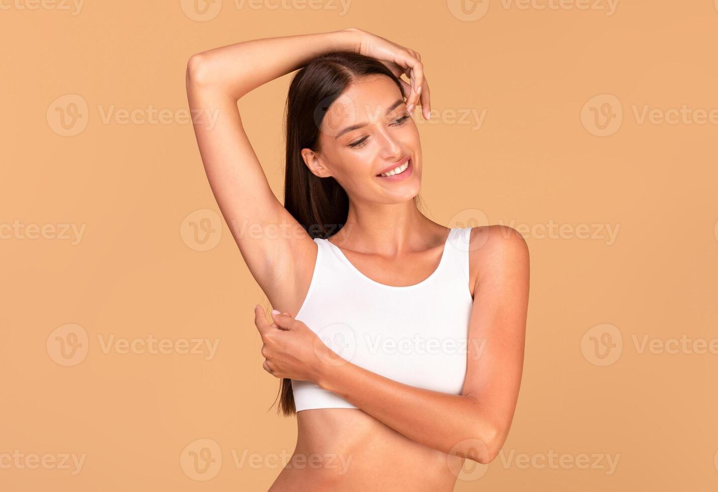Reducing body odor. Happy fit lady standing with one hand up, demonstrating her smooth depilated armpit, posing in white top over beige background photo