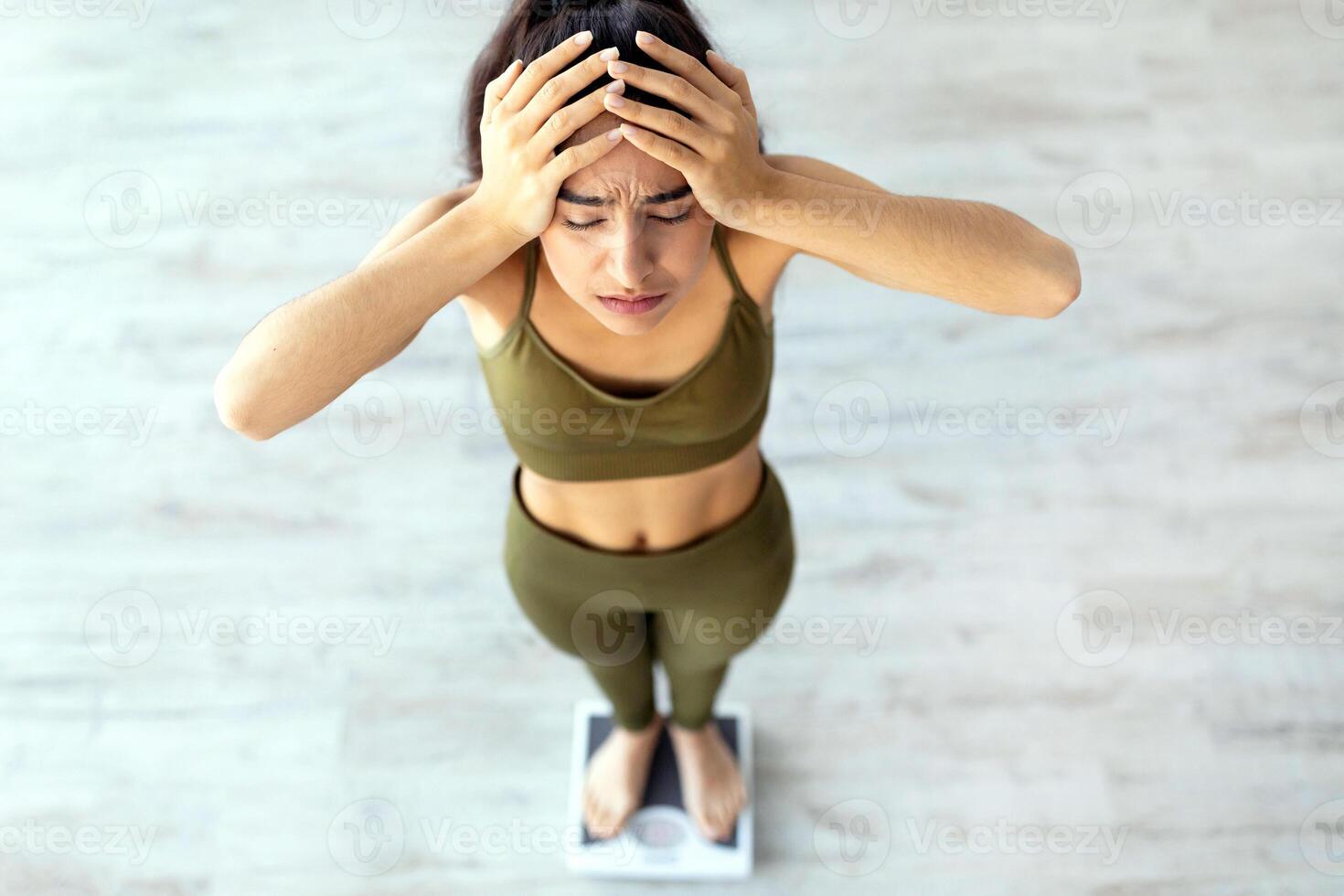 Unsuccessful slimming and dieting concept. Upset young Indian woman unhappy about her weight, standing on scales, holding head in disappointment indoors, above view photo