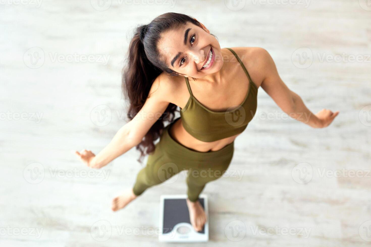 Above view of millennial Indian lady dancing on scales, choosing healthy lifestyle, happy with slimming result. Young Hindu woman celebrating her weight loss success photo