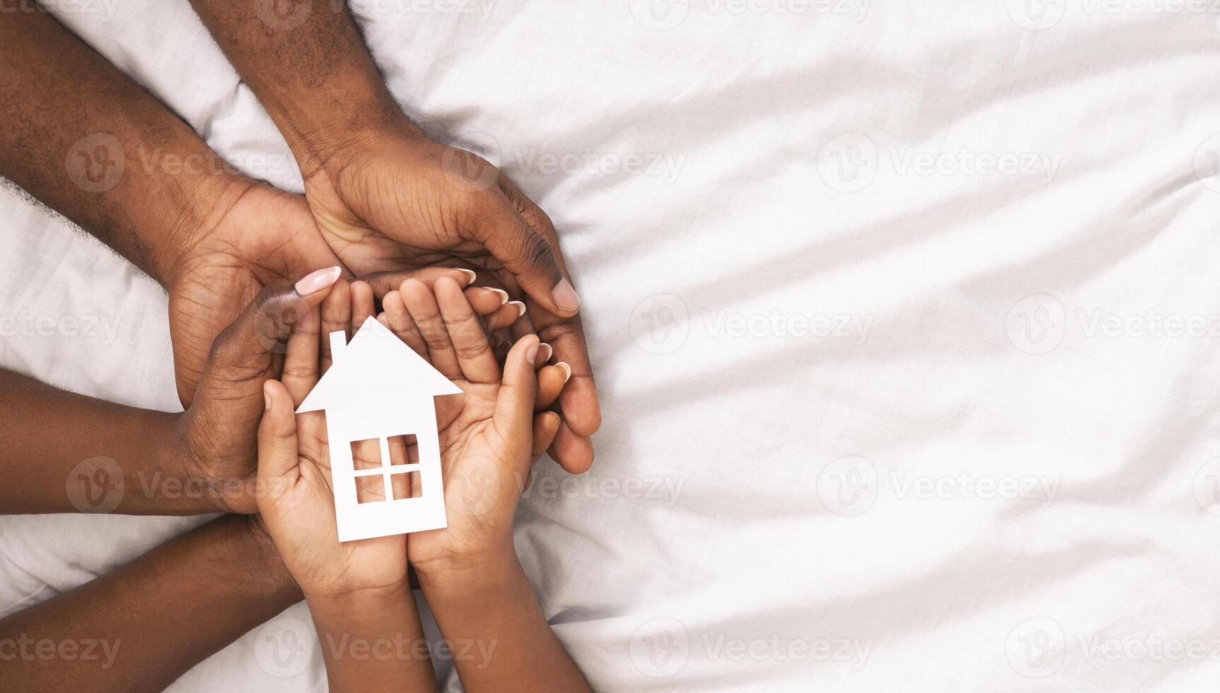Adoption concept. Black family of three holding paper house figure in hands over white bed sheet background, panorama with free space photo