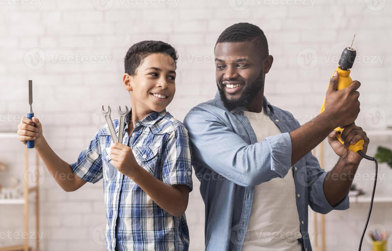 Father Son Activities. Portrait Of Cheerful Afro Boy And His Dad Standing Back To Back With Construction Tools In Hands photo