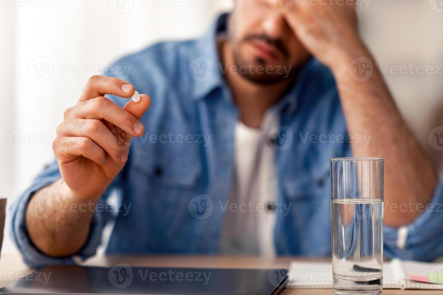 Exhausting Headache. Sick young millennial man taking medicine with glass of water, holding pill, suffering from migraine at home or at workplace. Selective focus on tablet, blurred background photo