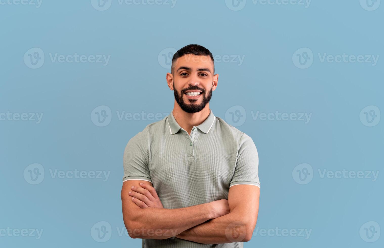 Happy arab guy smiling to camera, crossing hands and posing over blue background, studio shot. Excited self-confident middle eastern man expressing positive emotions photo