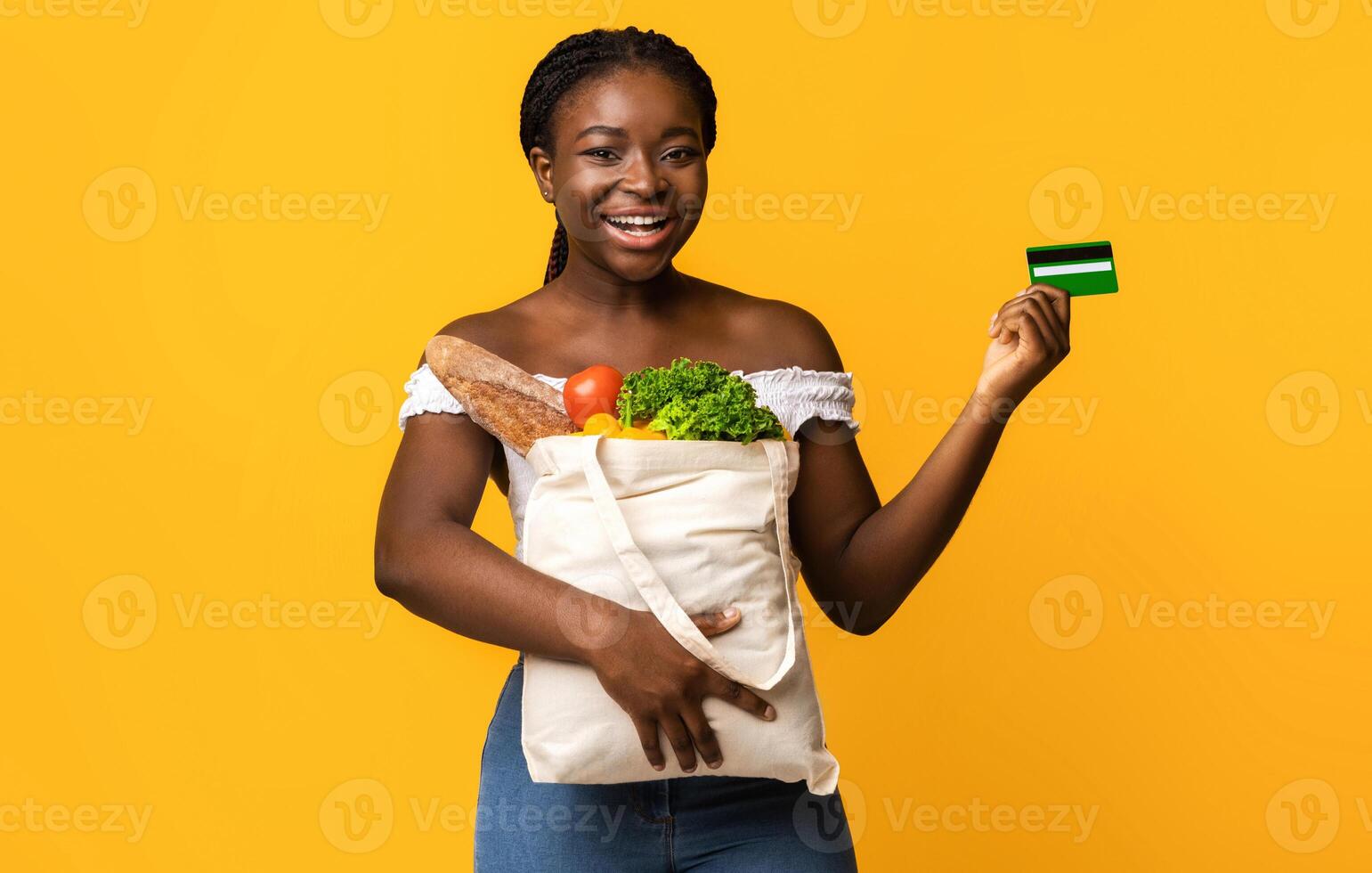 Happy black woman holding shopping bag with groceries and showing credit card, smiling at camera, cheerful african lady carrying package with fresh vegetables and fruits, isolated on yellow background photo