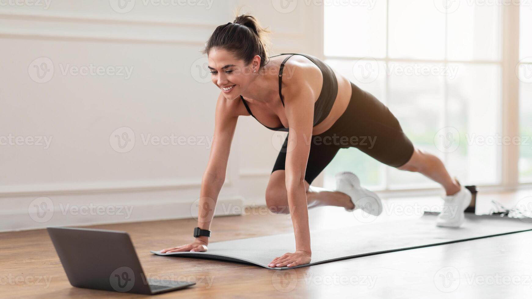 Morning Routine Concept. Cheerful slim lady in black sportswear and sneakers doing mountain climber exercise on yoga mat and watching online tutorial, looking at computer screen on floor at home gym photo