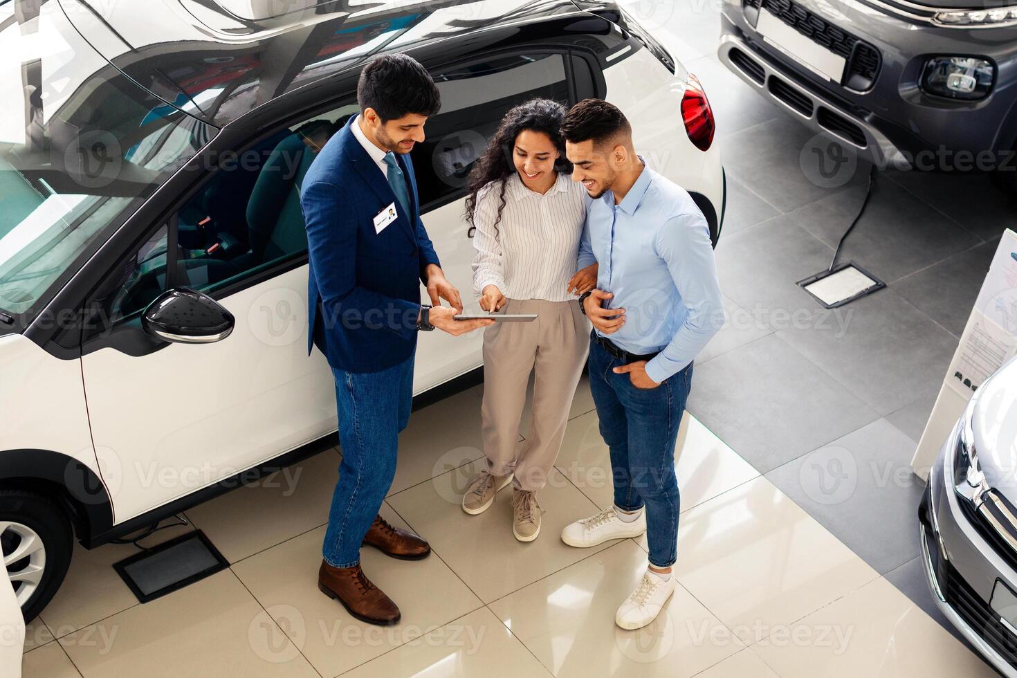 Top view of wealthy middle eastern young couple having conversation with male sales assistant at luxury auto showroom, checking nice white car and smiling, full length photo, copy space photo