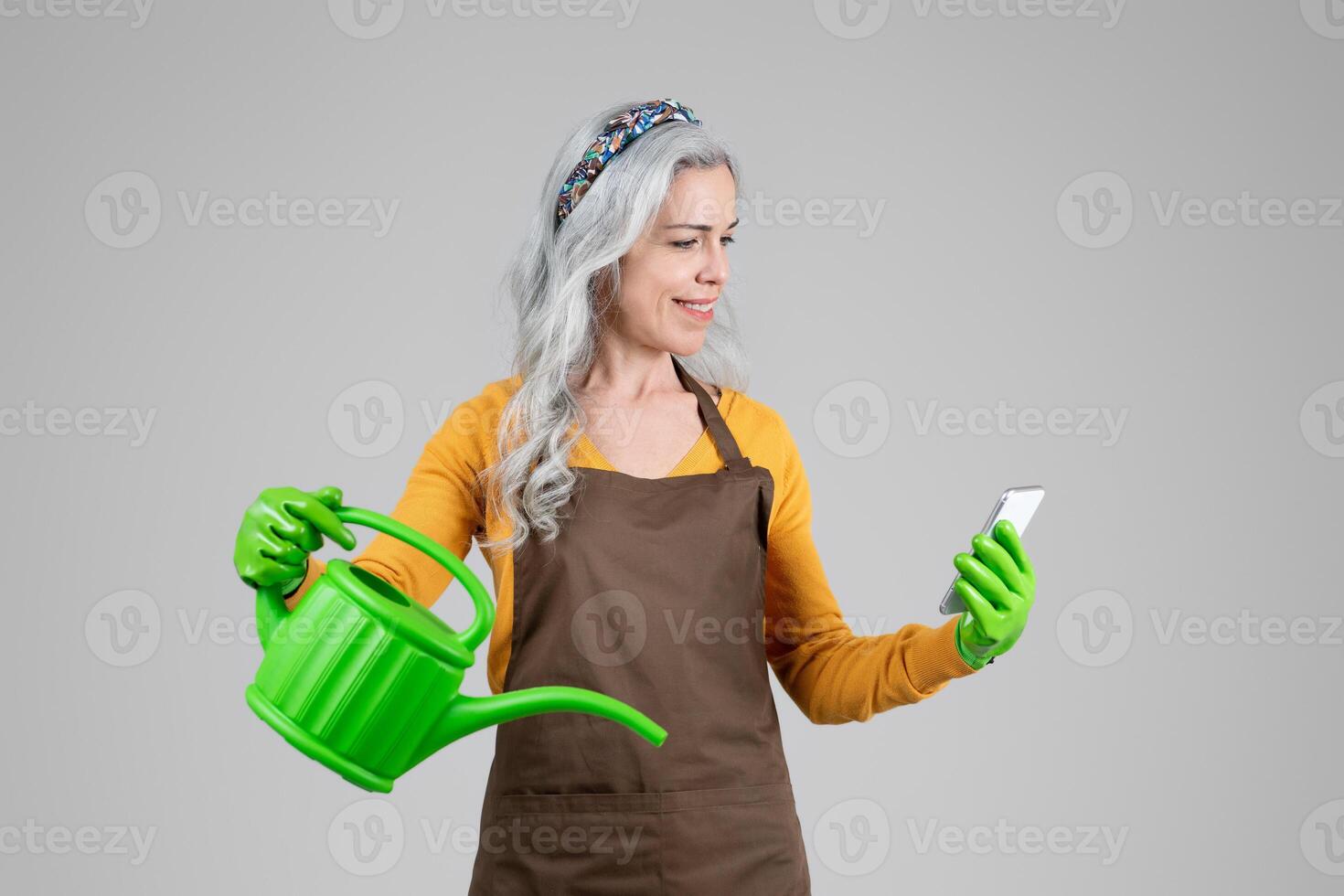 A middle aged woman dressed for gardening is smiling as she holds a green watering can in one hand and checks her phone with the other. Her gray hair is styled neatly, and she wears gloves. photo