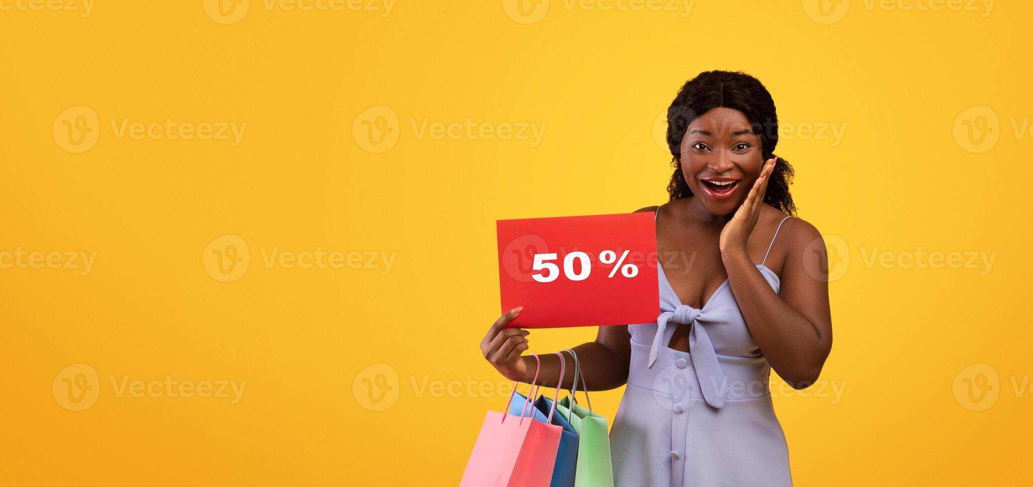 Seasonal sales. Emotional young black woman holding bright shopping bags and FIFTY PERCENT OFF sign, looking at camera in excitement, cannot believe huge discount over orange studio background photo