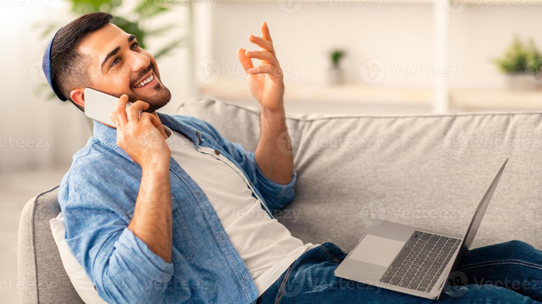 Portrait of cheerful excited millennial man talking on smart phone, gesturing using pc sitting on sofa. Happy young jewish male chatting with friends distracted from work taking break photo