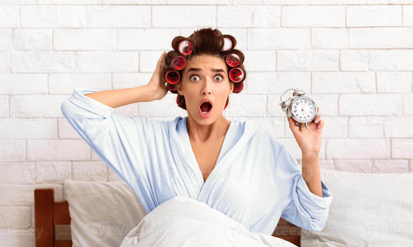 Oversleep Concept. Shocked Woman With Curlers Holding Clock Being Late For Work Sitting In Bed At Home In The Morning photo