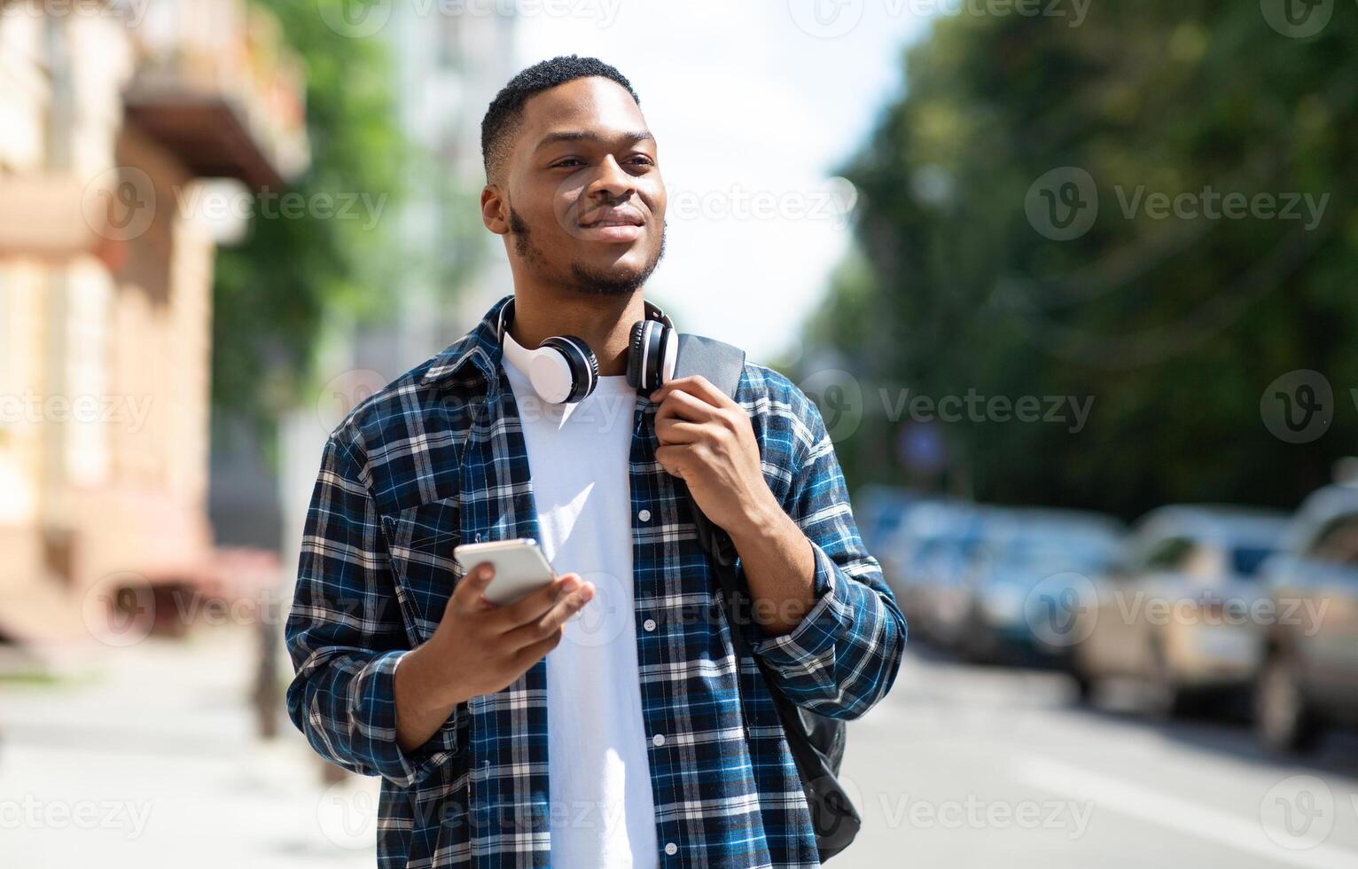 Front view portrait of black stylish guy walking on a sunny day with cell phone, wearing backpack and headset, copyspace photo