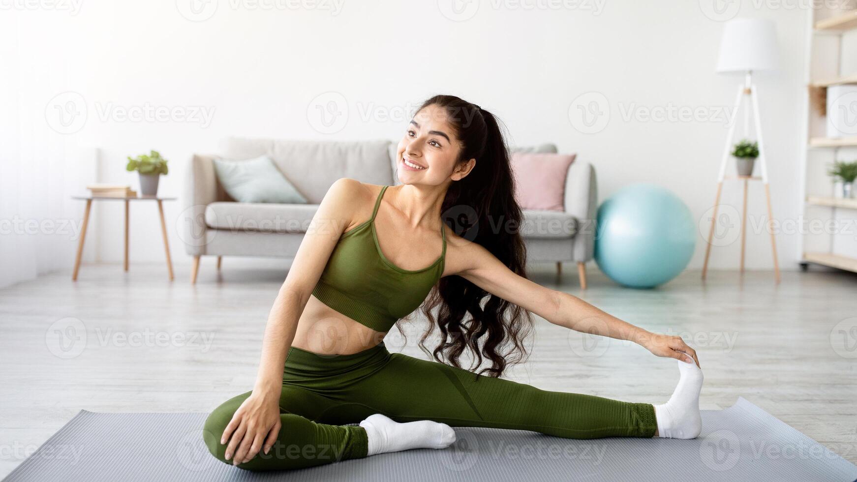 Domestic sports concept. Athletic Indian woman in sportswear doing fitness exercises on yoga mat, stretching her legs at home. Fit young Eastern lady staying flexible during covid lockdown photo