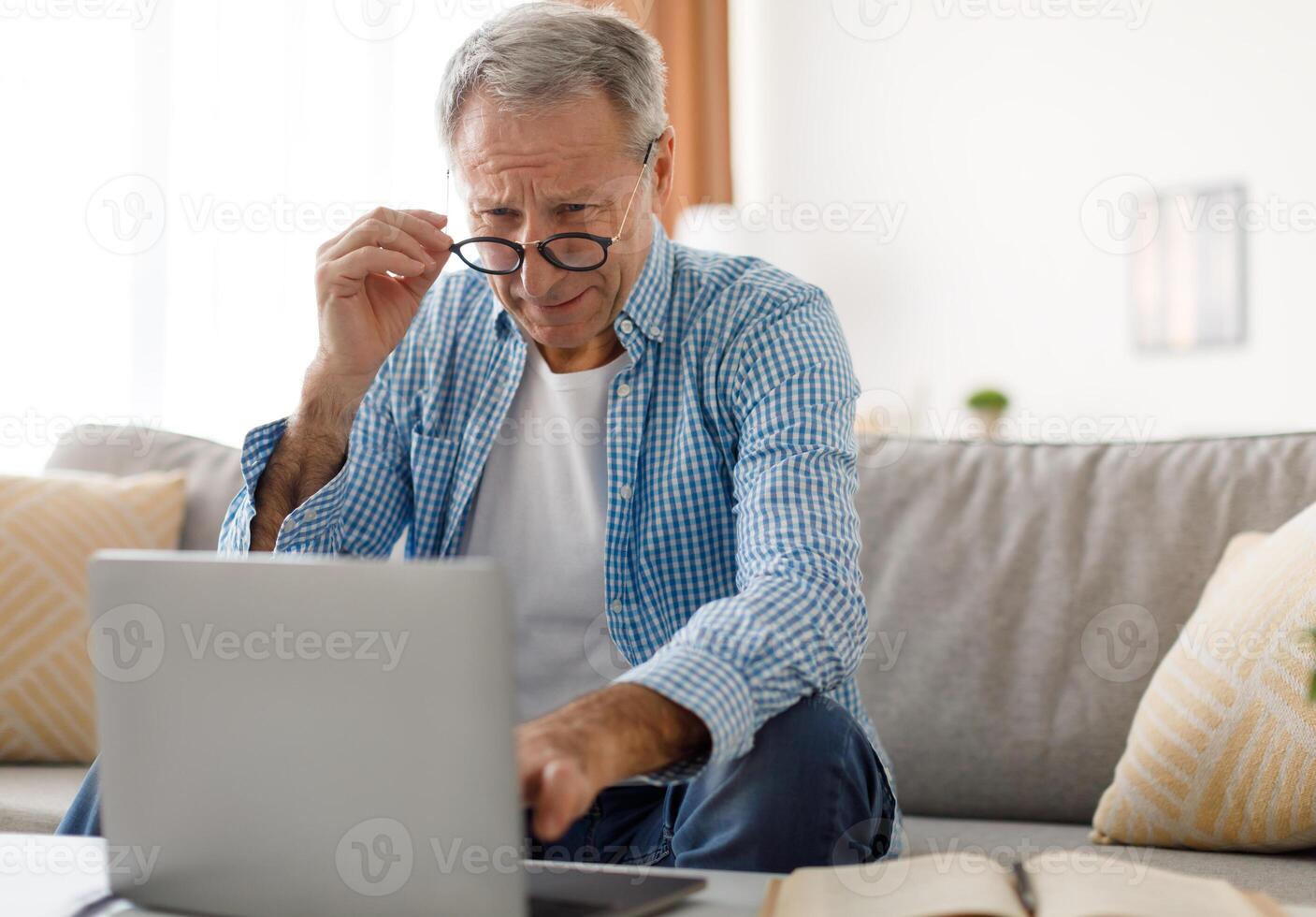 Poor Eyesight. Senior Man Squinting Eyes Using Laptop Wearing Eyeglasses Having Problems With Vision Sitting On Couch. Ophtalmic Issue, Bad Sight In Older Age, Macular Degeneration Concept photo