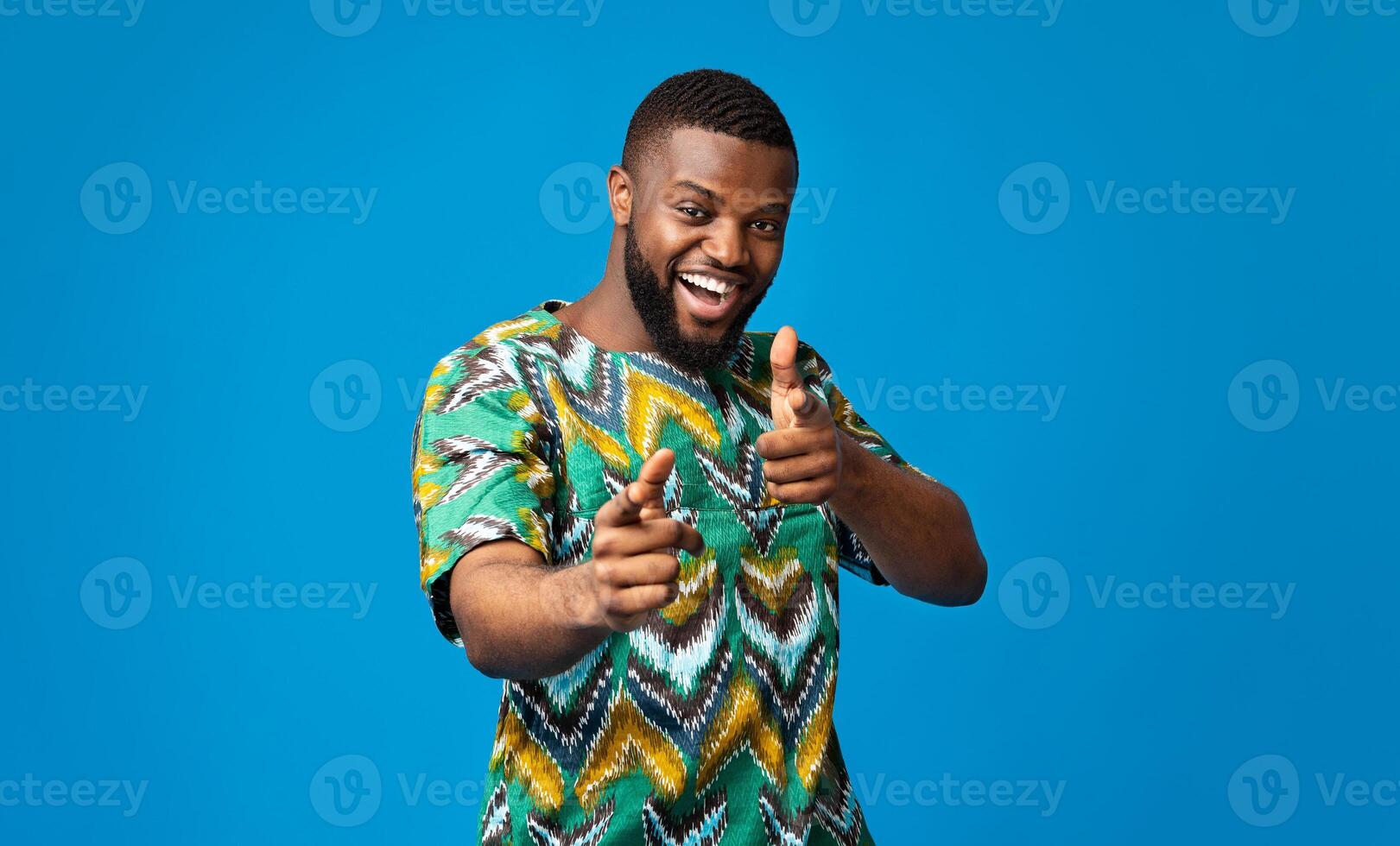 Cheerful black man in traditional african clothes indicating to camera over blue photo