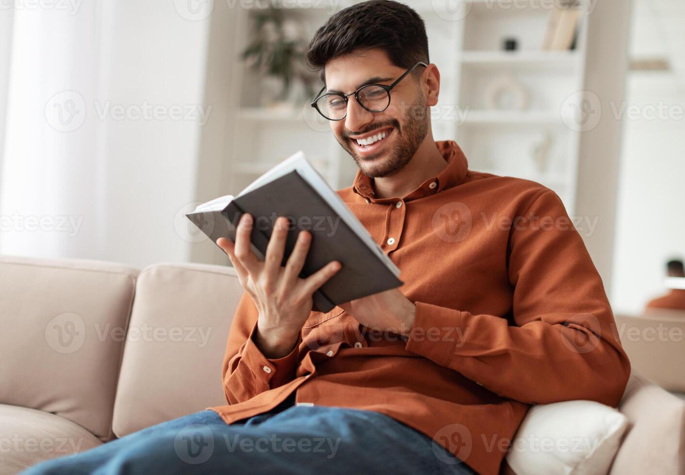Leisure and Relaxation Concept. Portrait of cheerful smiling young Middle Eastern man wearing eyeglasses holding paper book, sitting on couch at home in living room, reading literature, enjoying story photo