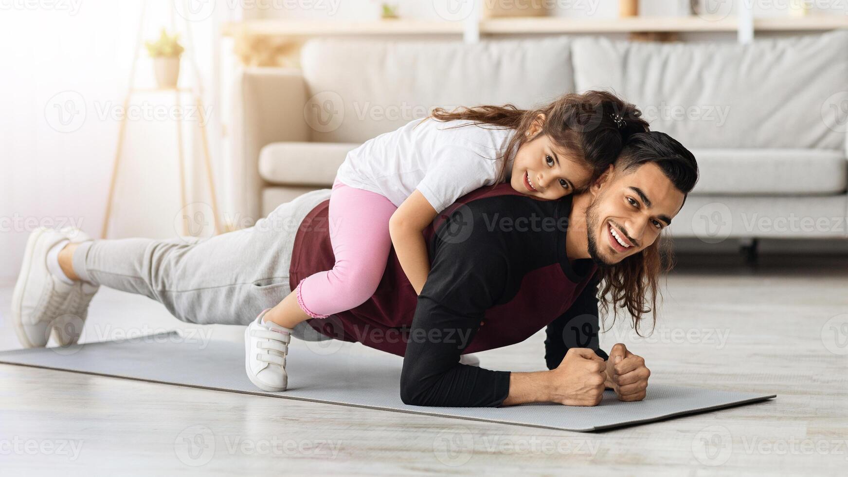Happy sporty middle-eastern father and kid bonding while training at home. Athletic man planking with little daughter on his back, cheerfully smiling at camera, living room interior, copy space photo