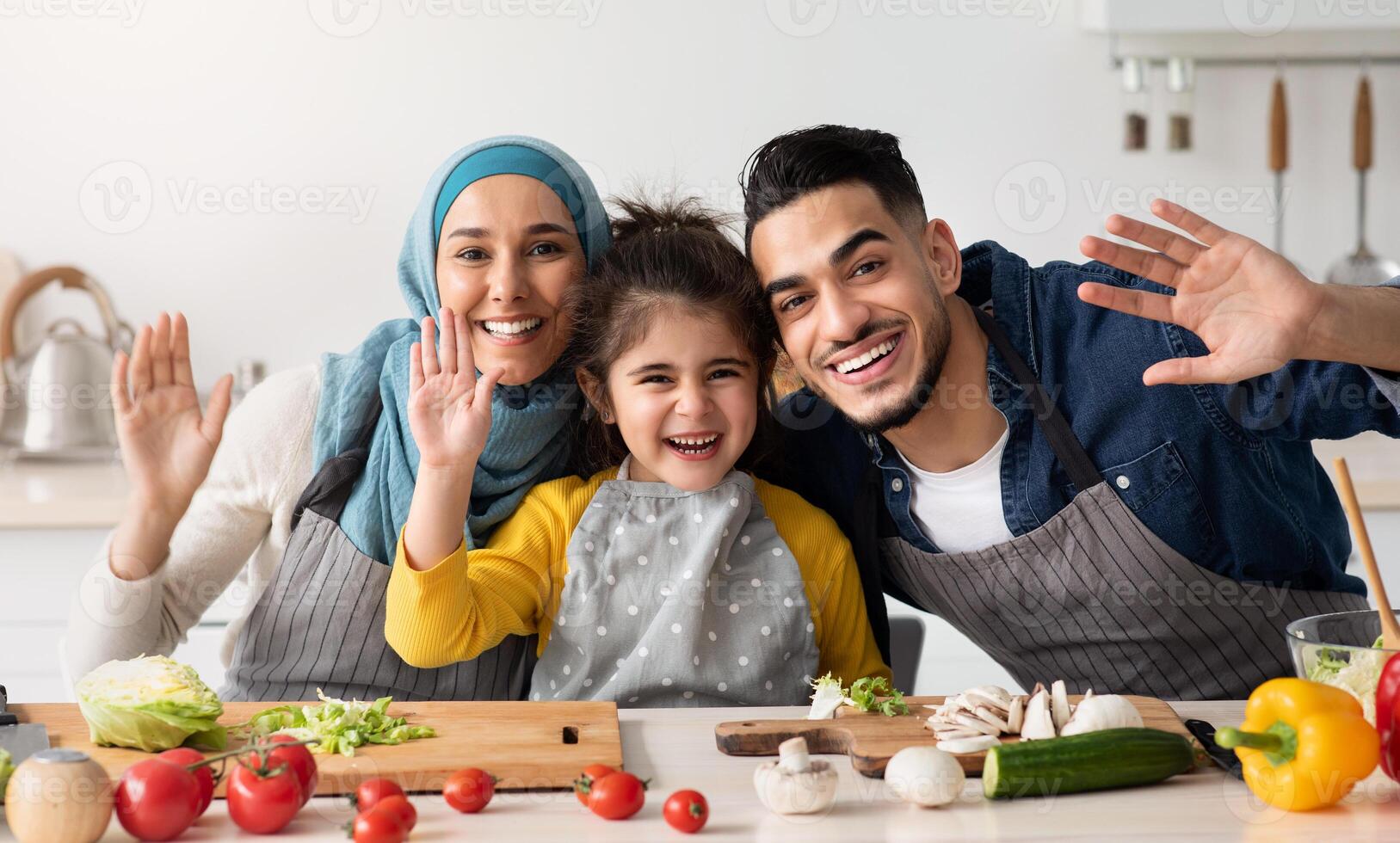 Cheerful Islamic Family Of Three Waving Hands At Camera While Cooking In Kitchen Together, Happy Middle Eastern Parents And Their Little Daughter Having Fun While Prepsring Food At Home, Free Space photo