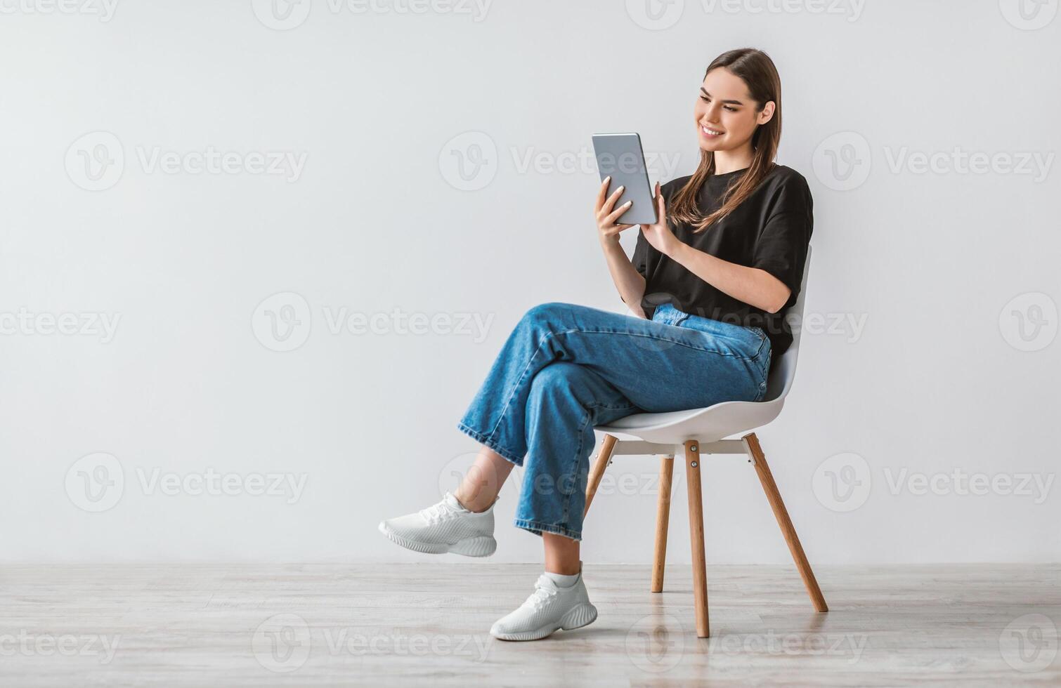 Full length portrait of cheerful young woman with digital tablet sitting on chair, working online against white studio wall, free space. Modern technologies and communications concept photo