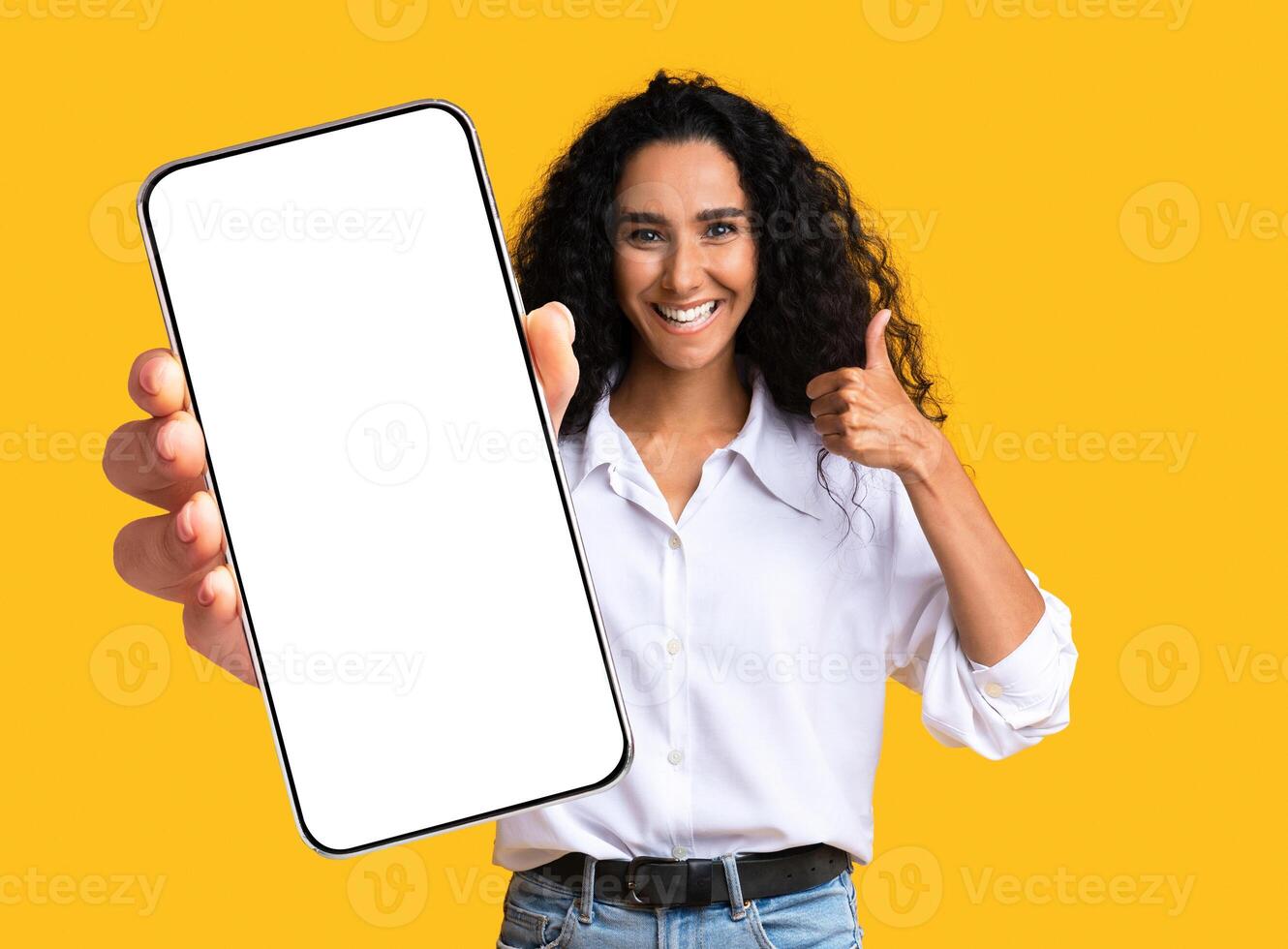 Cute pretty young middle-eastern woman showing modern mobile phone with empty screen and thumb up, closeup of cellphone with blank display, mockup, shot on yellow studio background, copy space photo