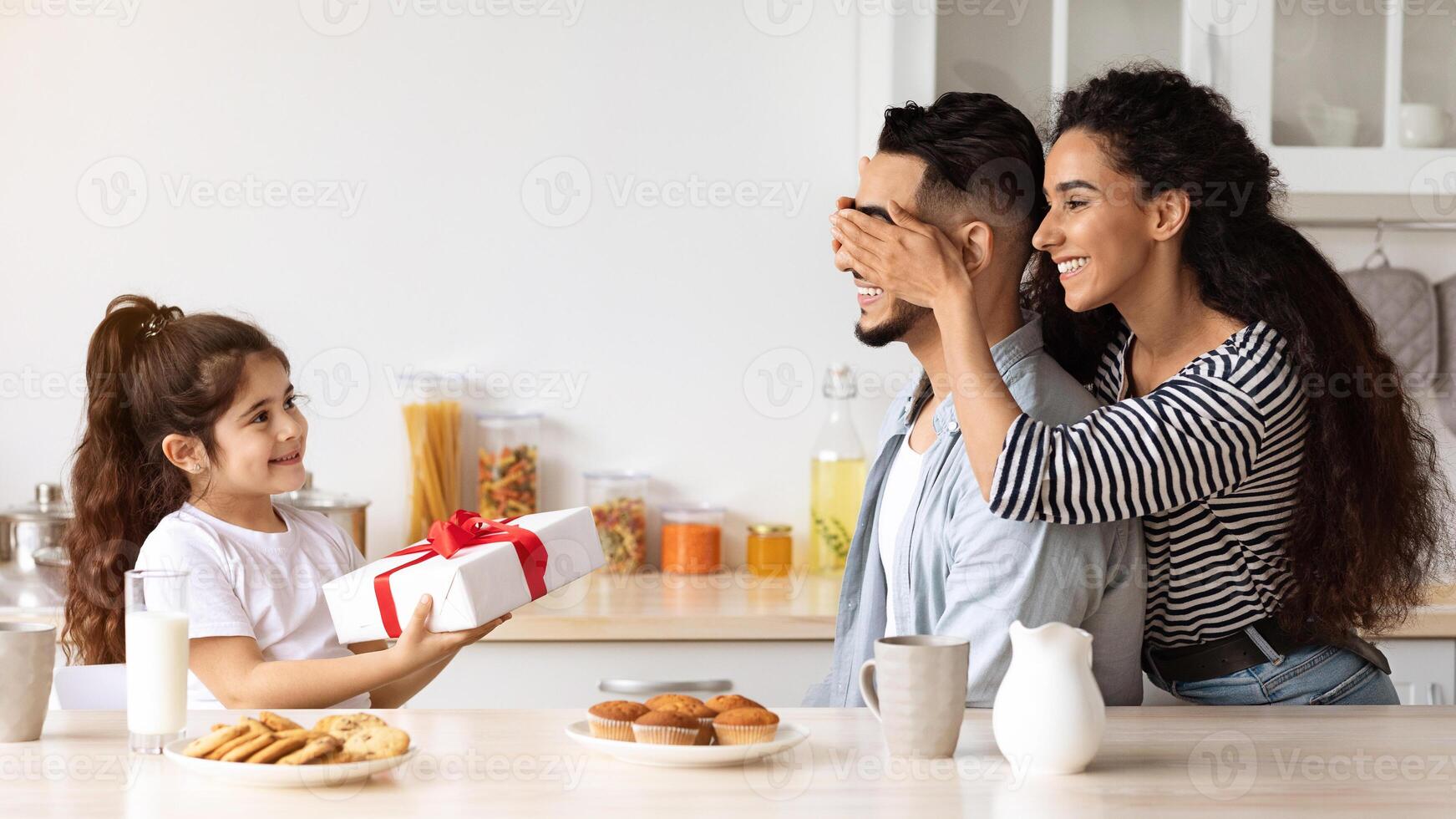 Loving middle-eastern mother and daughter greeting their father with birthday, closing his eyes and giving big festive gift box while having breakfast together in kitchen. Family birthday celebration photo