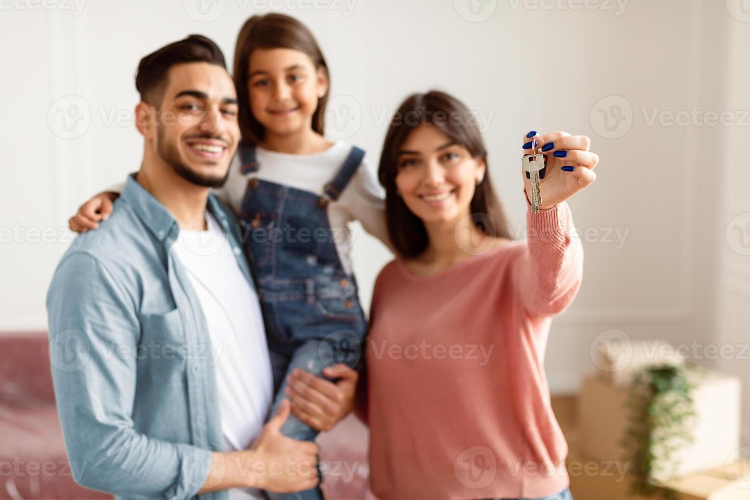 Positive family of three showing house key, standing in living room of new home, selective focus hand. Parents and daughter happy to move to bigger apartment, holding flat key, blurred background photo