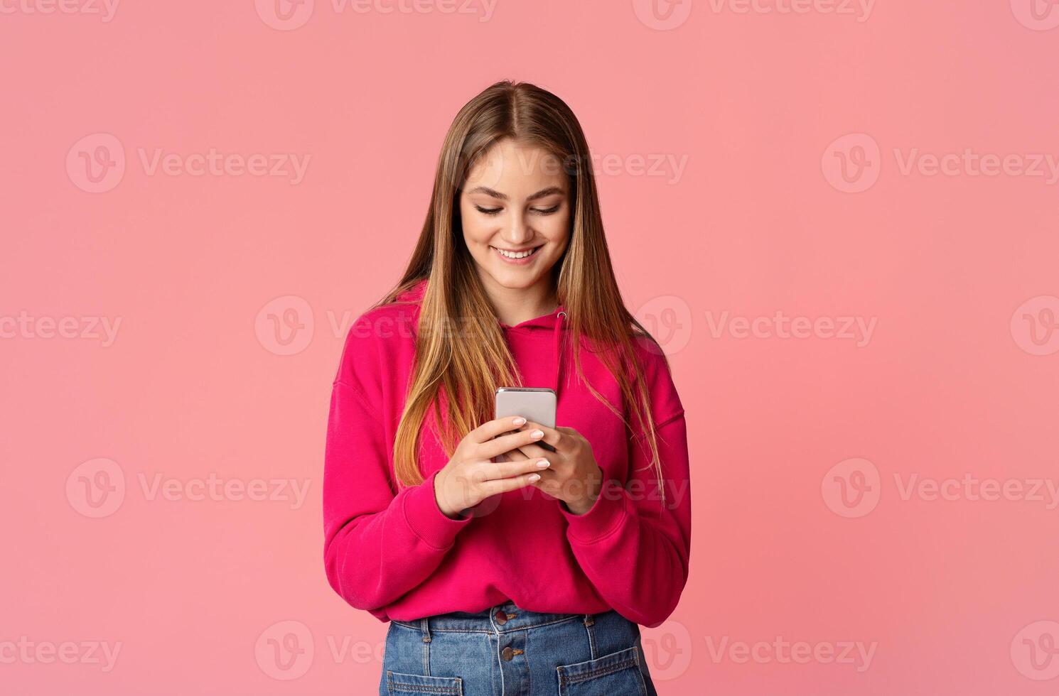 Good news in message. Smiling teenage girl checking smartphone isolated on pink background, studio shot photo