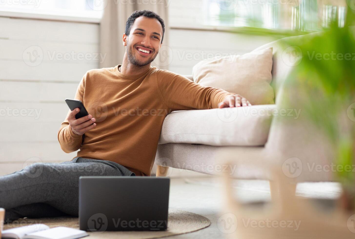 Freelance Career. Portrait Of Excited Middle Eastern Male Holding Cell Phone Working On Pc At Home Office, Happy Guy Using Computer Sitting On Floor Leaning On Sofa In Living Room, Looking Away photo