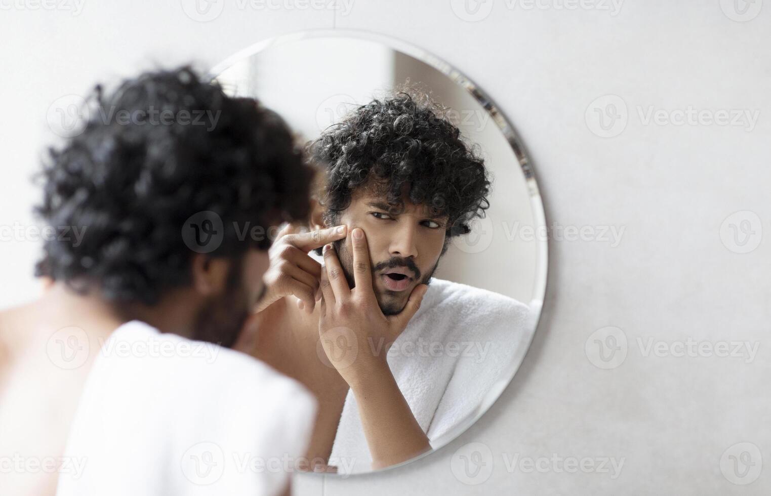 Problem skin. Concerned indian man popping pimple on cheek while standing near mirror in bathroom. Unhappy guy inspecting face, suffering acne, focus on reflection photo