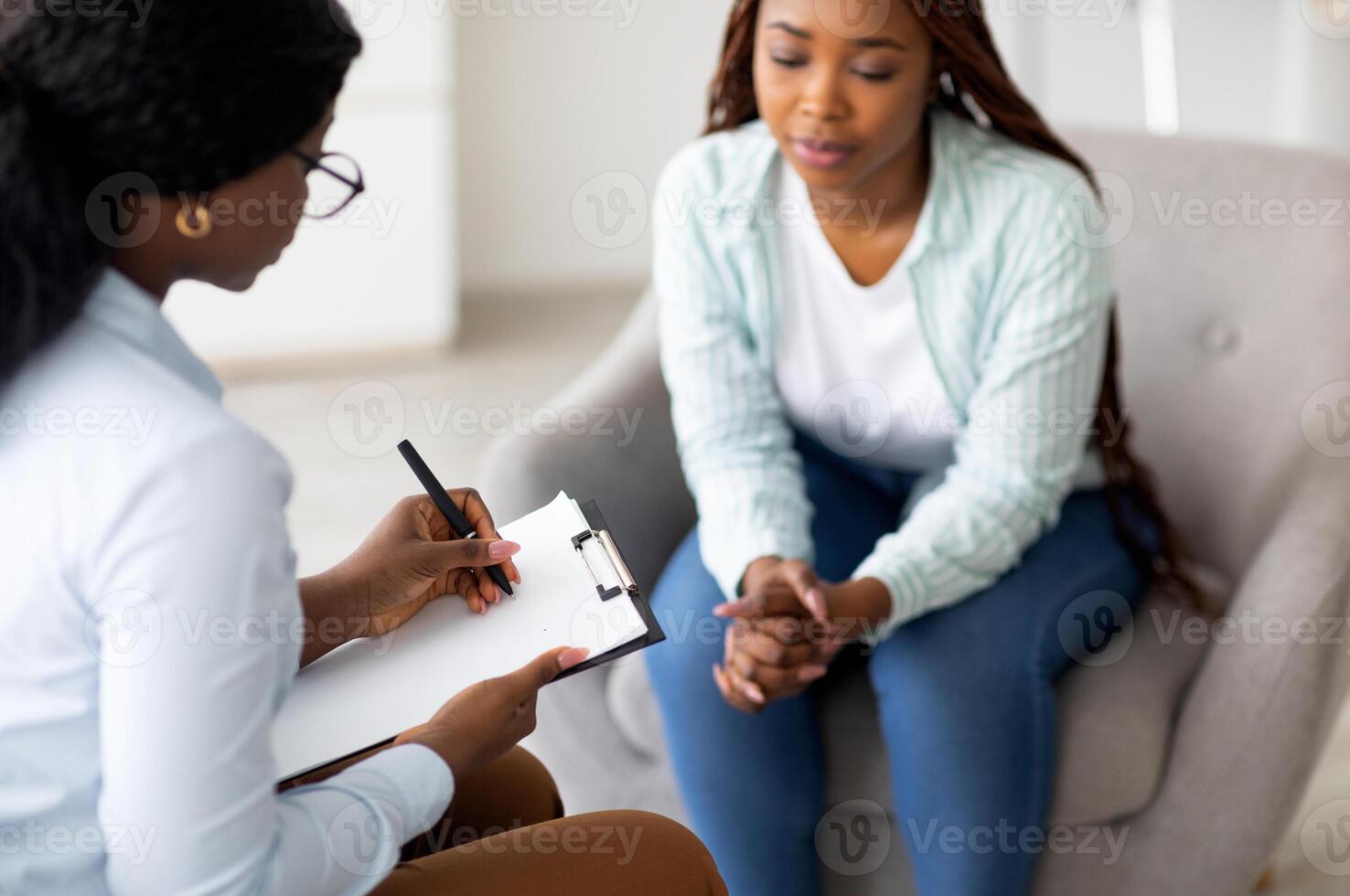 Stressed black female patient consulting psychologist at medical office, selective focus. Psychiatrist helping sad African American woman with nervous problems, taking notes during session photo