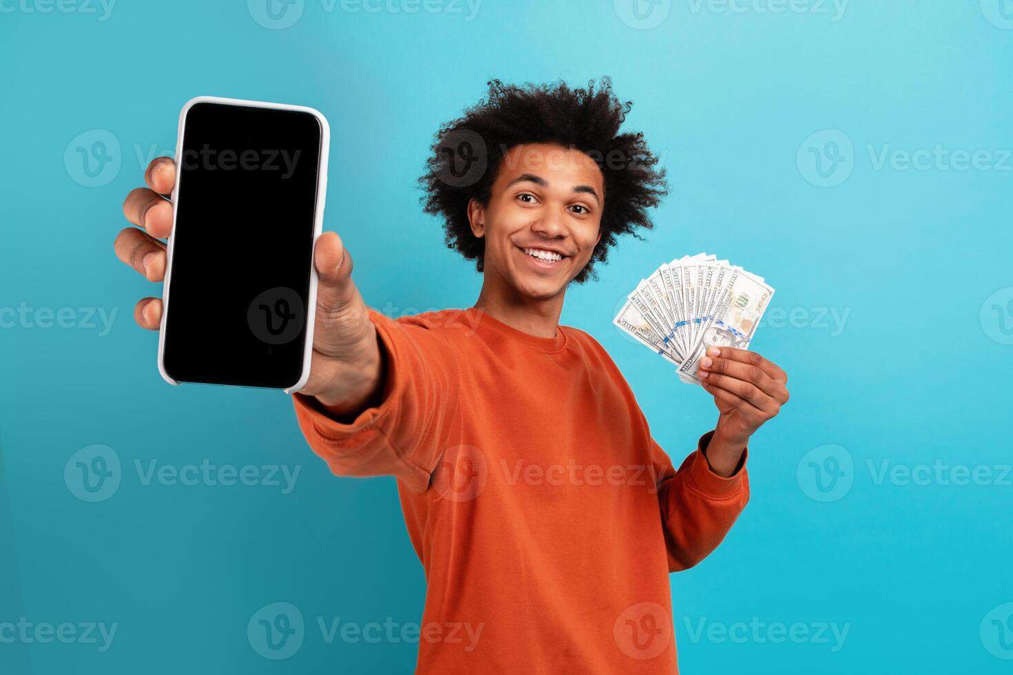 A young African American man with curly hair smiles widely while holding a smartphone in one hand and cash in the other. He stands in front of a bright blue background, exuding joy and confidence. photo