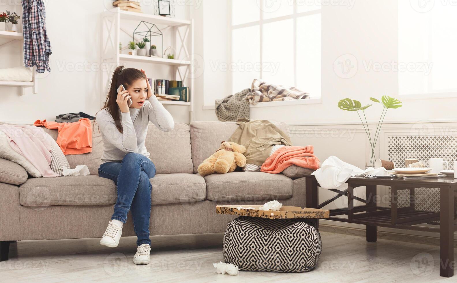 Desperate helpless woman sitting on sofa in messy living room. and talking on mobile, surrounded by many stack of clothes. Disorder and mess at home, copy space photo