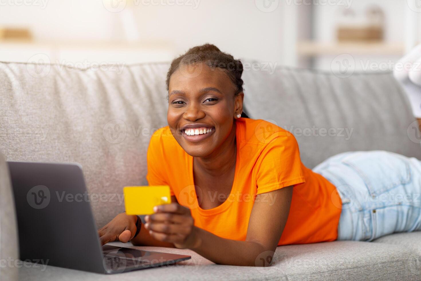 Joyful Afro woman shopping online with laptop pc and credit card, lying on couch at home. Millennial black lady searching web for goods on dicsount, buying at internet store, ordering remotely photo