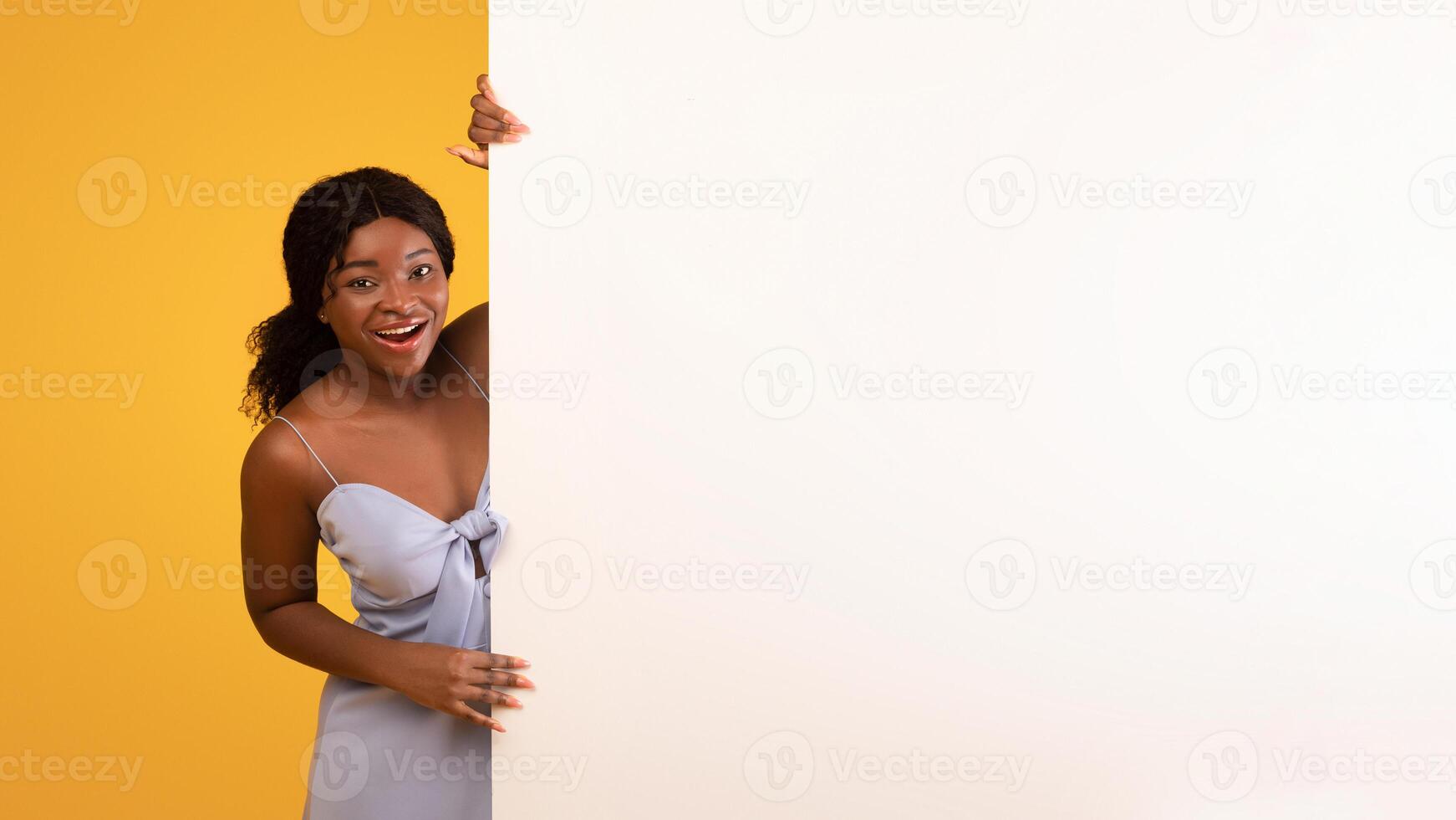 Smiling young black lady looking out from behind blank white paper banner with space for ad on orange studio background. Millennial African American female offering mockup for advertisement design photo