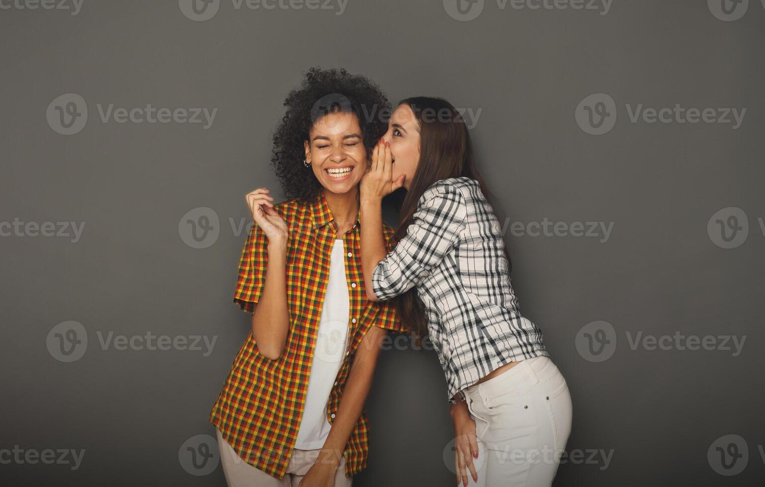 Young woman telling her girlfriend some secret. Two women gossiping. Excited emotional girl whispering to her friend ear, gray studio background photo