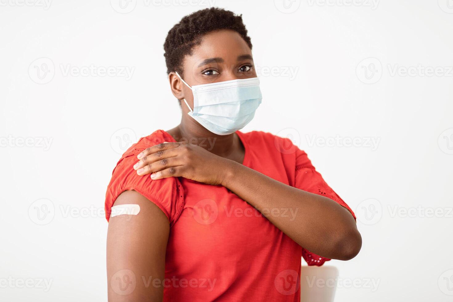 I'm Vaccinated. Black Woman Showing Arm With Plaster Strip After Getting Covid-19 Coronavirus Vaccine Injection Posing On White Studio Background, Wearing Protective Face Mask. Covid Vaccination photo